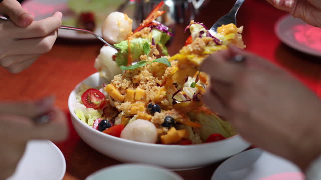 A cropped view of an Asian family enjoying and sharing a bowl of mixed fruits and vegetables salad together at a restaurant