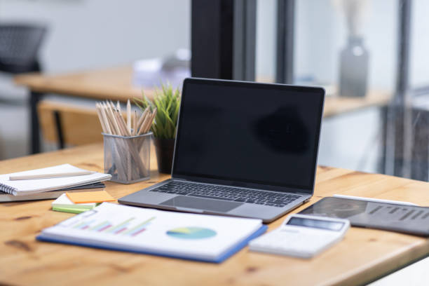 istock Laptop Computer finance graph, notebook, and eyeglasses sitting on a desk in a large open plan office space after working hours 1584353811
