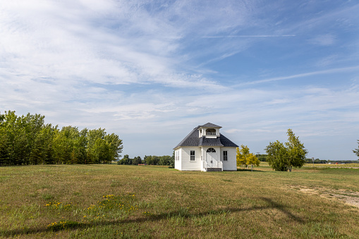 This image shows a summer landscape view of a rural 19th century wood constructed one-room country schoolhouse situated on a prairie in midwestern USA, with blue sky background.