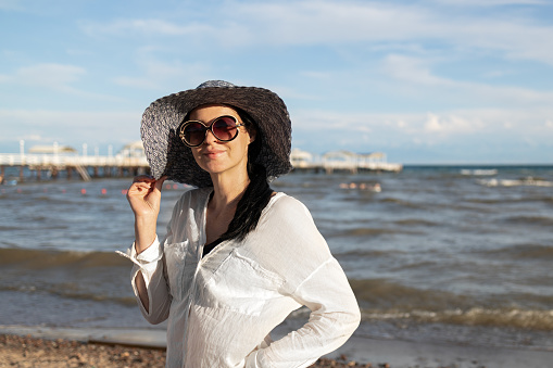 Smiling brunette woman wearing a white shirt, dark hat and sunglasses walking along the beach at stormy weather.