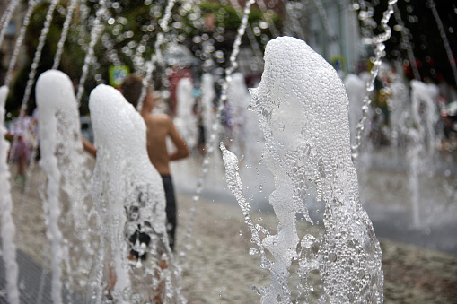 Under jets of city's pedestrian fountain, shooting from ground, children run around playing on hot summer day. Children's entertainment run along pedestrian fountain, close up foaming jet water
