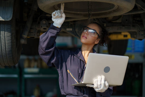 african american mechanic woman using laptop computer checking list to undercarriage of car in workshop at auto car repair service center with lift . car engineer female inspection vehicle details stock photo