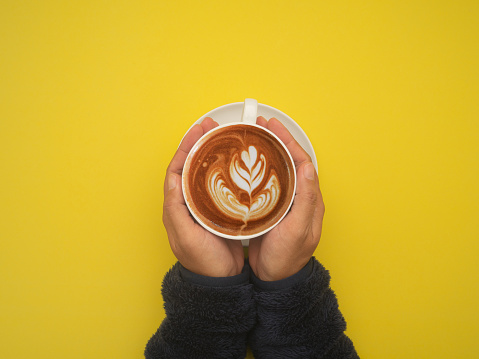 Top view of hands woman holding a fresh coffee cup over a yellow background. Space for text. Beverage and relaxation concept