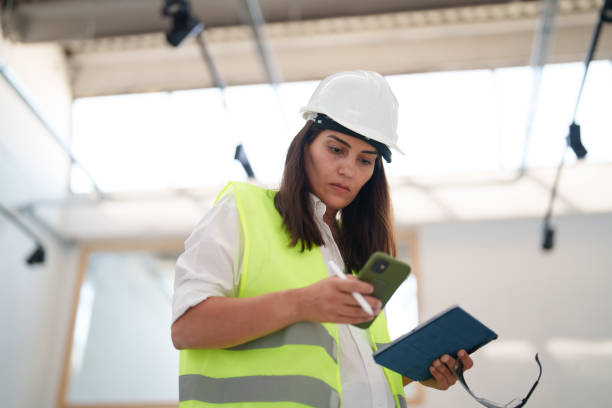 istock Woman wearing protective white helmet, using mobile phone at the construction site 1584657094