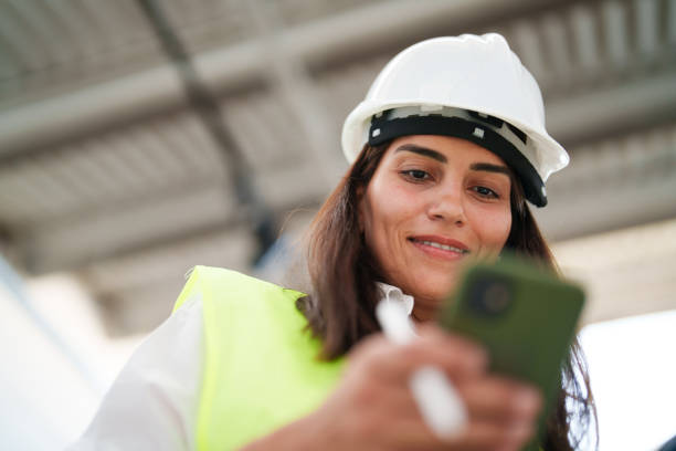 istock Woman wearing protective white helmet, using mobile phone at the construction site 1584657143