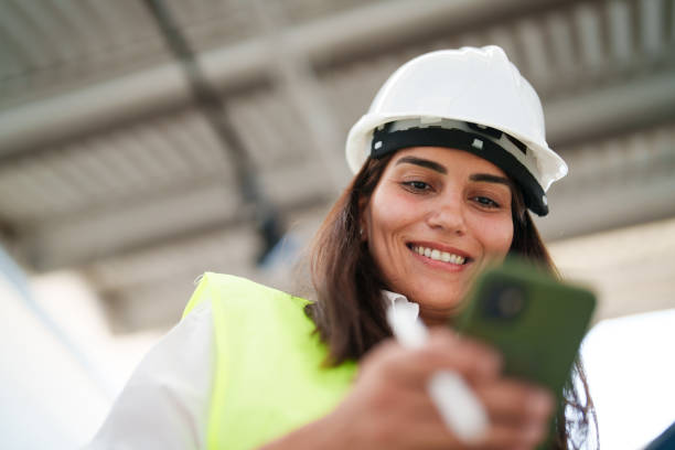 istock Woman wearing protective white helmet, using mobile phone at the construction site 1584658195