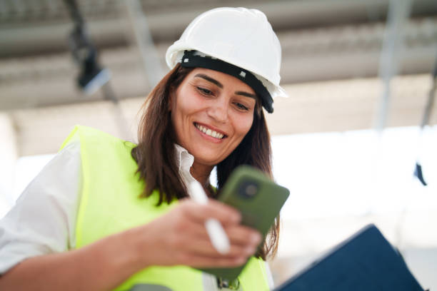 woman wearing protective white helmet, using mobile phone at the construction site - female construction telephone building contractor imagens e fotografias de stock