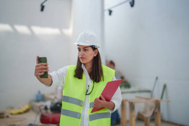 istock Woman wearing protective white helmet, using mobile phone at the construction site 1584664737