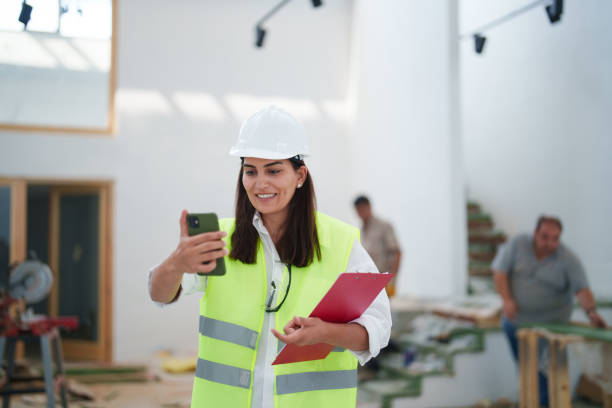 istock Woman wearing protective white helmet, using mobile phone at the construction site 1584664742