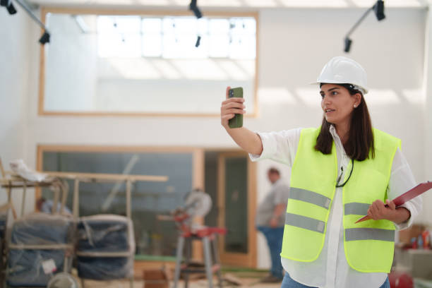 istock Woman wearing protective white helmet, using mobile phone at the construction site 1584666443