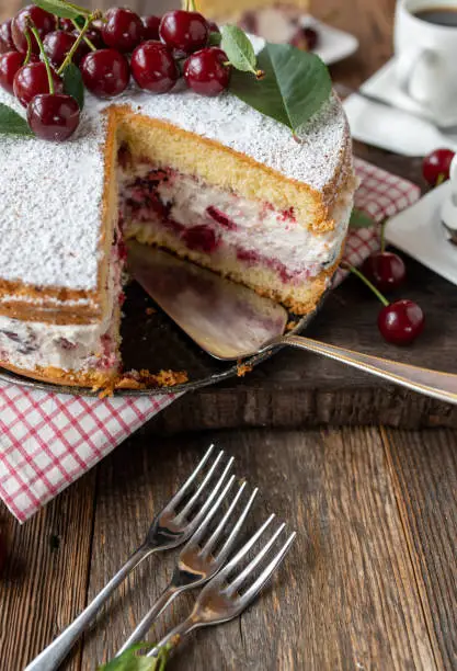 Homemade  baked cream cake with fresh sour cherries. Served with cross section view and whole on rustic and wooden table background with forks and coffee.