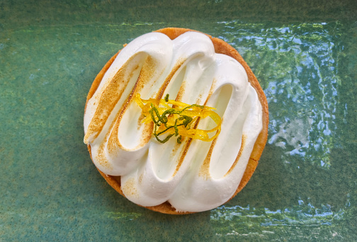 Mini lemon pies with meringue, lemon peel, lemon leaves, whisks with meringue, a fork, and some piping nozzles, on a white background with copy space.