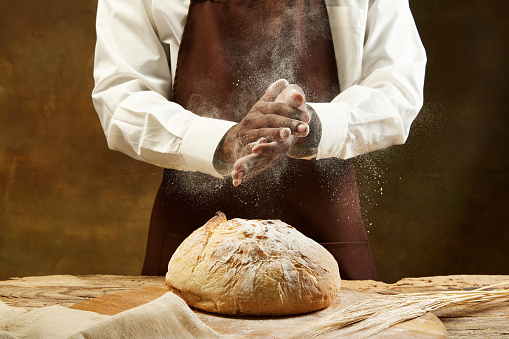 Unrecognizable baker in bround apron pouring flour on round loaf of bread over brown background. Concept of food, health, traditions, bakery and cooking. Ad