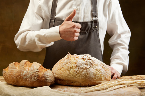 Unrecognizable baker in grey apron likes round bread and long loaf over brown background. Concept of food, health, traditions, bakery and cooking. Ad