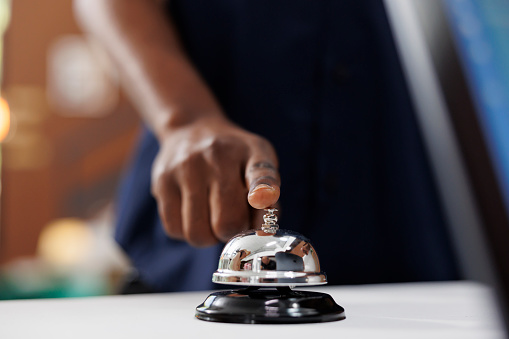 Impatient hotel guest using service bell at reception counter calling for receptionist to provide check-in forms. Close-up shot of african american person ringing concierge bell at front desk.