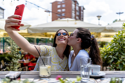 Embracing the magic of the moment, two female friends seize the opportunity for a selfie, documenting and celebrating their beautiful bond amidst the tranquil and picturesque setting of the cafe garden