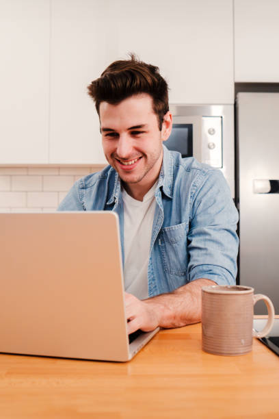 istock Vertical portrait of handsome man working with a laptop computer at home kitchen drinking coffee at the morning. Happy young entrepreneur or student searching information on internet. Freelance work 1584825133