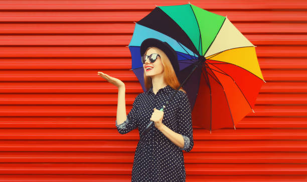 Autumn portrait of stylish happy smiling young woman holding colorful umbrella wearing black round hat on red background stock photo