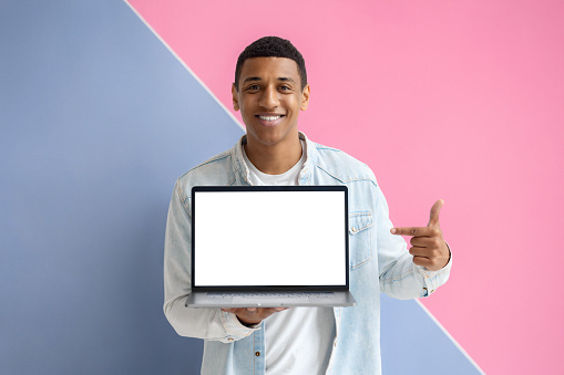Young African American man with laptop on blue background. Male points her finger at a blank laptop screen, looking at the camera and smiling
