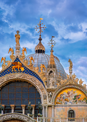 Details. Main entrance decorations, St Mark's or San Marco cathedral, Venice. Italy. UNESCO World Heritage city, blue sky, clouds. Rich decorations in mosaics and marble carvings, St Mark statue, Lion of St Mark bas-relief, two horses. Vertical shot