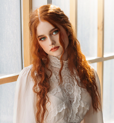Red-haired woman in vintage dress stands at large classic window waiting love. Clothing costume countess old style white blouse, brown long skirt Curly red hair hairstyle. Redhead girl princess 1800s stylish gown beauty face waiting love
