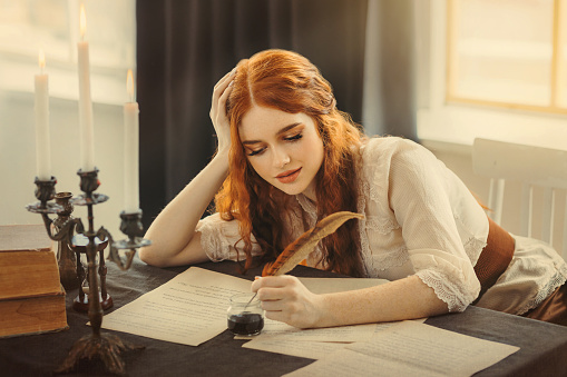 Medieval red-haired beauty woman writer holds pen feather quill in hands, sits at table writes letter on sheet paper. Vintage dress ancient room antique candlestick, old books. Red hair Girl smiling student studying.