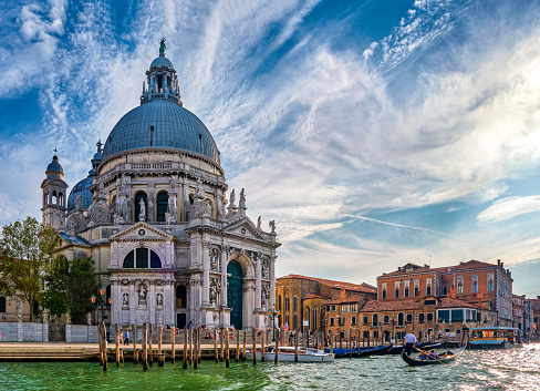 Beautiful view of iconic basilica di Santa Maria della Salute or St Mary of Health and colorful palaces or palazzo by waterfront of Grand Canal, Venice, Italy. Landmark on waterway by St Mark square, gondola pass by, blue sky, daylight, great clouds.