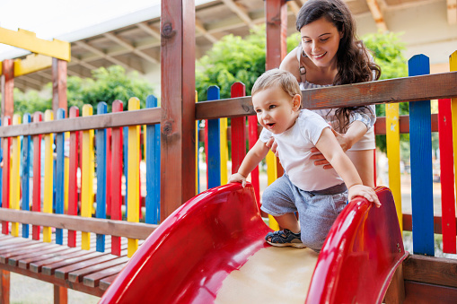 Beautiful young mother and cute baby boy having fun playing on a slide in the park, mother helping him climb on the top