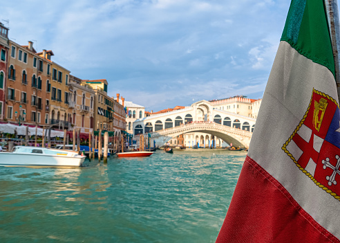 Beautiful view of Italian maritime flag and bridge of Rialto or ponte Rialto on Grand Canal on Venice, Italy. Gondola service station on the left. UNESCO world heritage city. Daylight, sunshine, soft clouds. Selective focus.