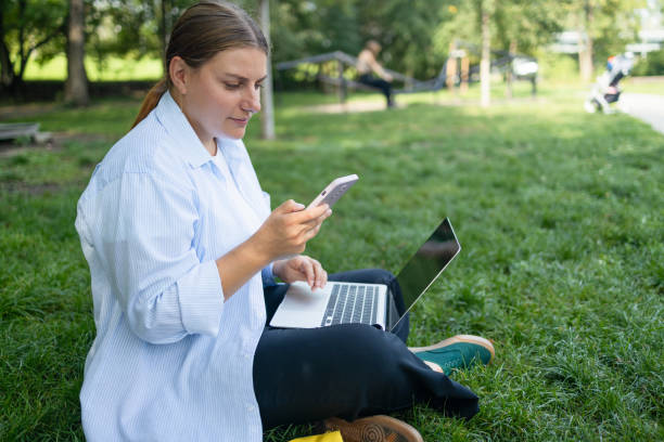 istock Happy Caucasian student freelancer woman rest use laptop pc computer talk by mobile cell phone look, sitting on grass outdoors in spring park. She is keyboarding on laptop. Freelance work concept. 1584873552