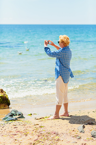 Elderly woman standing on the beach by the sea and taking photo of the sea and seagulls on mobile phone. Active seniors concept