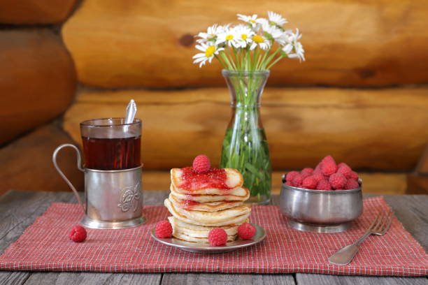 On a wooden table there is a stack of pancakes with fresh raspberries and jam near a bowl of ripe berries, a glass of tea and a vase of flowers.