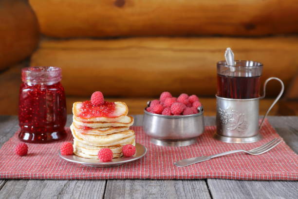 On a wooden table there is a stack of pancakes with fresh raspberries and jam near a bowl of ripe berries, a glass of tea and a glass jar of jam.