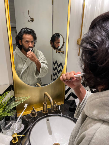 istock Image reflection in mirror of Indian man stood at bathroom sink wearing bathrobe cleaning teeth, toothpaste and manual toothbrush, foaming mouth, dental care, artificial fern plant in metal pot on marble bathroom counter, self care and grooming concept 1585002103