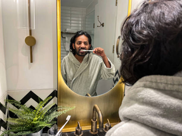 istock Image reflection in mirror of Indian man stood at bathroom sink wearing bathrobe cleaning teeth, toothpaste and manual toothbrush, foaming mouth, dental care, artificial fern plant in metal pot on marble bathroom counter, self care and grooming concept 1585002105