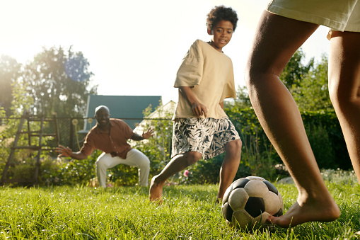 Legs of barefoot girl kicking soccer ball to her brother while playing on green lawn with him and their father standing on background