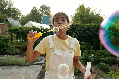 Youthful African American girl in coveralls and t-shirt blowing soap bubbles in front of camera while playing on backyard of country house