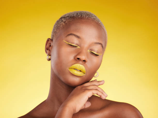 istock Face, makeup and lipstick with a black woman in studio on a yellow background for creative, beauty or cosmetics. Gold, color and eyeshadow on young model with eyes closed for fashion or aesthetic 1585038445