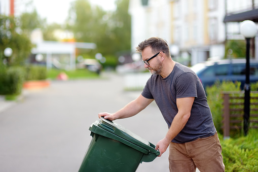 Mature man pulling out a large green plastic garbage container in front of the townhouse to the roadway of the street. Garbage collection and recycling trash, the zero waste concept. Waste management