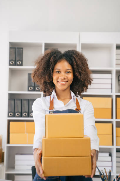 istock African American female holding parcels and looking to camera, standing among several boxes in modern office. 1585120406