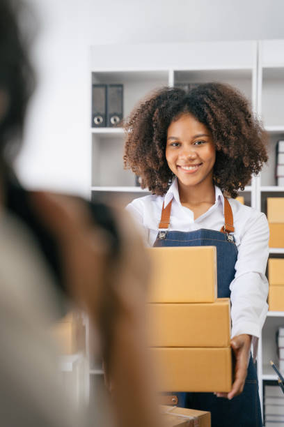 istock African American female holding parcels and looking to camera, standing among several boxes in modern office. 1585120541