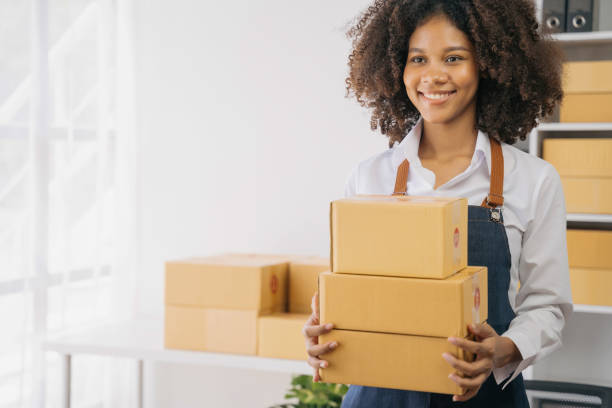 istock African American female holding parcels and looking to camera, standing among several boxes in modern office. 1585120546