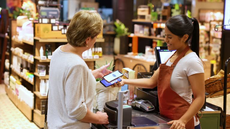 Friendly cashier holding a smartphone while mature female customer scans the code and makes a payment for her groceries at the supermarket