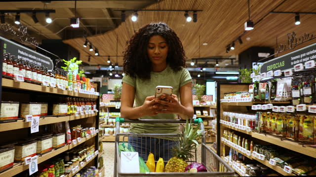 Black young woman leaning on the shopping cart while walking through the supermarket and checking her shopping list on smartphone
