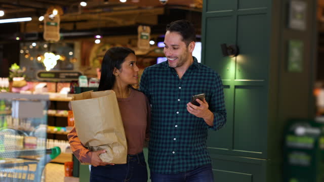 Happy Latin American couple leaving the supermarket carrying a shopping bag and holding a cell phone while talking and smiling