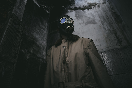 A man wearing a gas mask inside a ruined building with a leaky roof in the dark, apocalypse, good for book cover, low angle