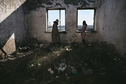 Two people, a guy and a girl in gas masks, stand inside an abandoned house next to the window and look back, apocalypse
