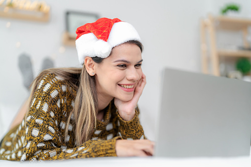 Beautiful woman lies comfortably on her bed, surrounded by the enchanting ambiance of Christmas decorations. Wearing cheerful Santa hat and exudes happiness while engaging in modern holiday activities using her laptop and wireless technology. Combining the pleasure of relaxation and enjoyment with long holiday.