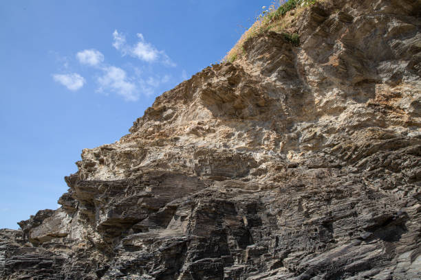 The top edge of a cliff in Cornwall viewed from underneath.  The sediment on the very top protrudes out and overhangs the actual cliff wall.\nEngland's coastline is being eroded as a result of storm surges and strong waves.  Prolonged wet weather can waterlog coastal areas and cause cliff collapse and rockfall. This process has been well documented by satellite imagery.  Some research suggests that the speed of coastal erosion will be increased by sea-level rises and more intense and frequent episodes of harsh weather.