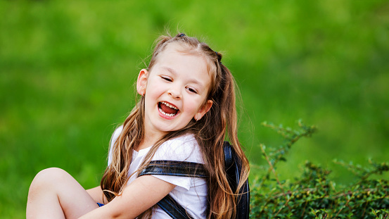 Adorable little girl is sitting on the grass and laughing. Little happy schoolgirl, education concept. Beginning of the academic year.Copy space.